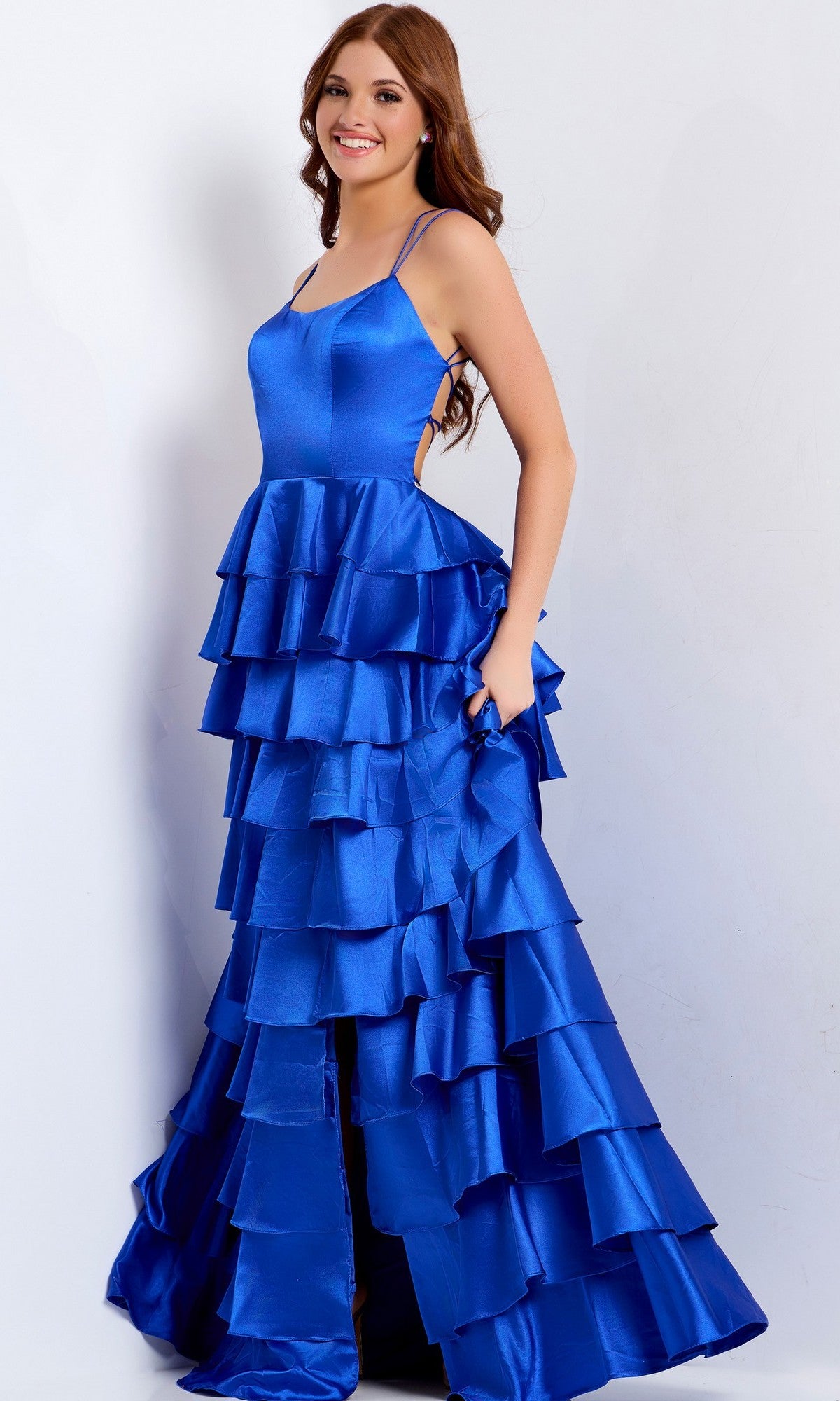 A woman models the Long Prom Dress: Jovani 48295, a sleeveless, royal blue gown with tiered ruffles and a strappy lace-up back, standing against a plain light background.